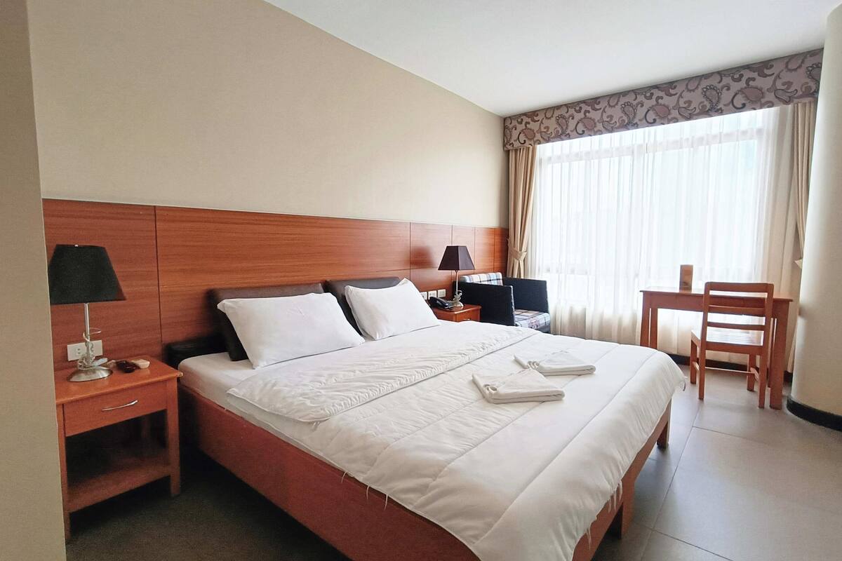 A hotel room with a large bed, a wooden headboard, a desk with a lamp, a chair, and a window with curtains.