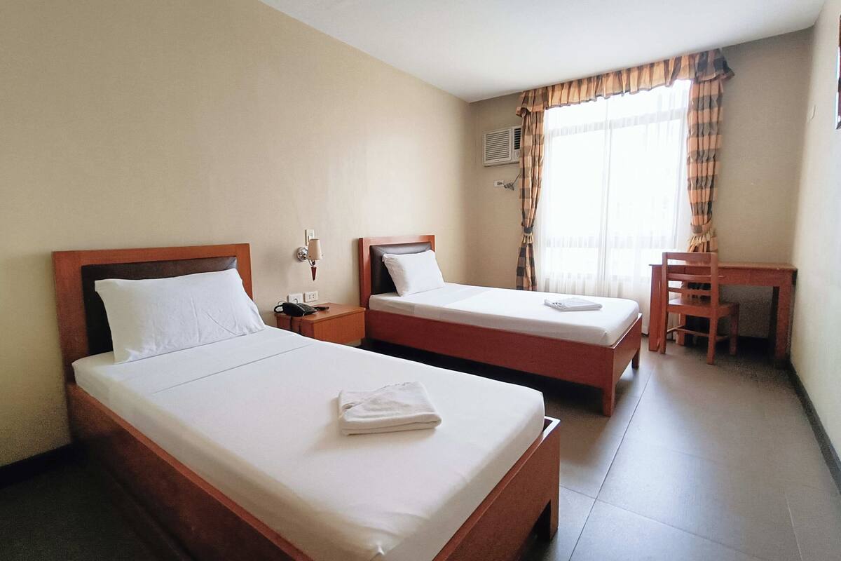 A hotel room with two single beds, a desk, a chair, and a window with curtains.