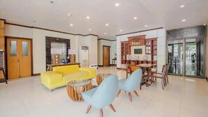 Un hall d’entrée spacieux avec un canapé jaune, un coin repas avec une table et des chaises en bois, et un comptoir de réception avec une enseigne.