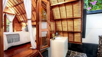 A bedroom with a wooden ceiling, a bed with a white canopy, a small bathroom with a sink and a mirror, and a stone floor.