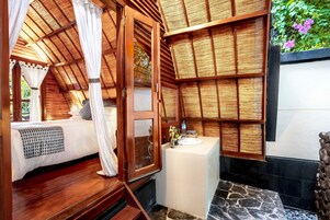 A bedroom with a wooden ceiling, a bed with a white canopy, a small bathroom with a sink and a mirror, and a stone floor.