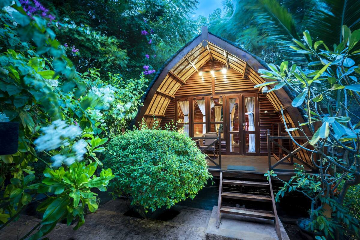Una cabaña de madera con techo inclinado, rodeada de vegetación exuberante y una pequeña piscina.