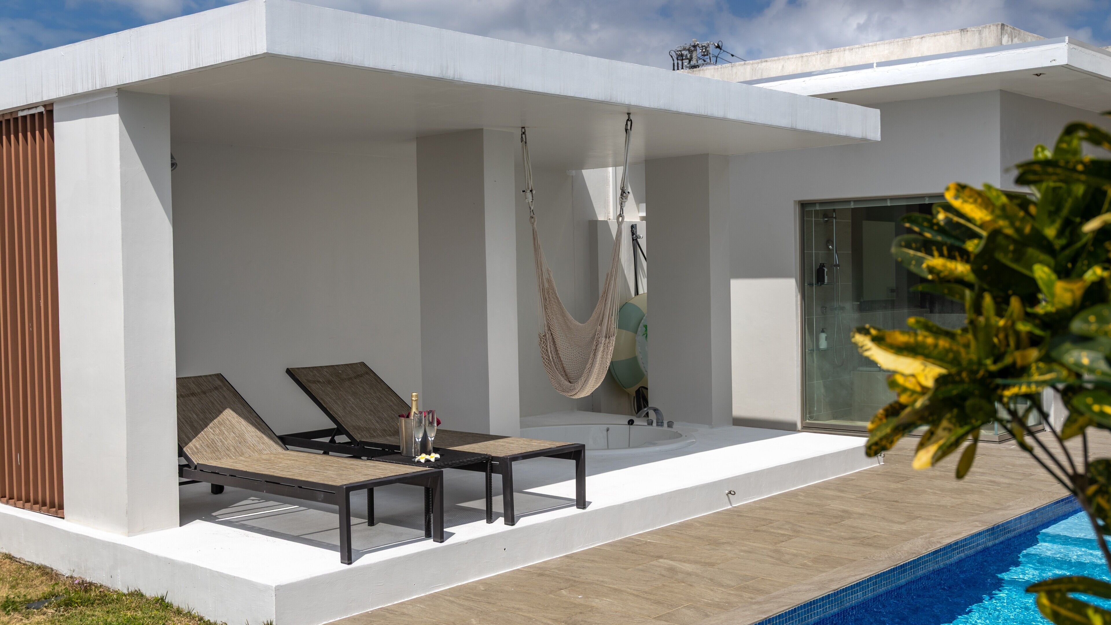 Un área de piscina exterior moderna con una hamaca, sillones reclinables y un vestuario de vidrio.
