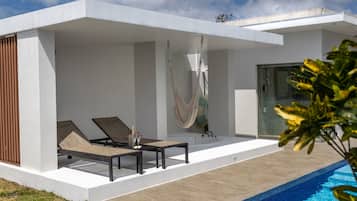 Un área de piscina exterior moderna con una hamaca, sillones reclinables y un vestuario de vidrio.