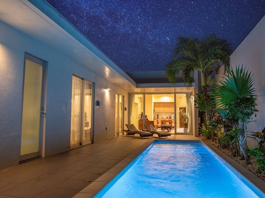 A swimming pool with lounge chairs, a covered patio, and a view of the night sky.