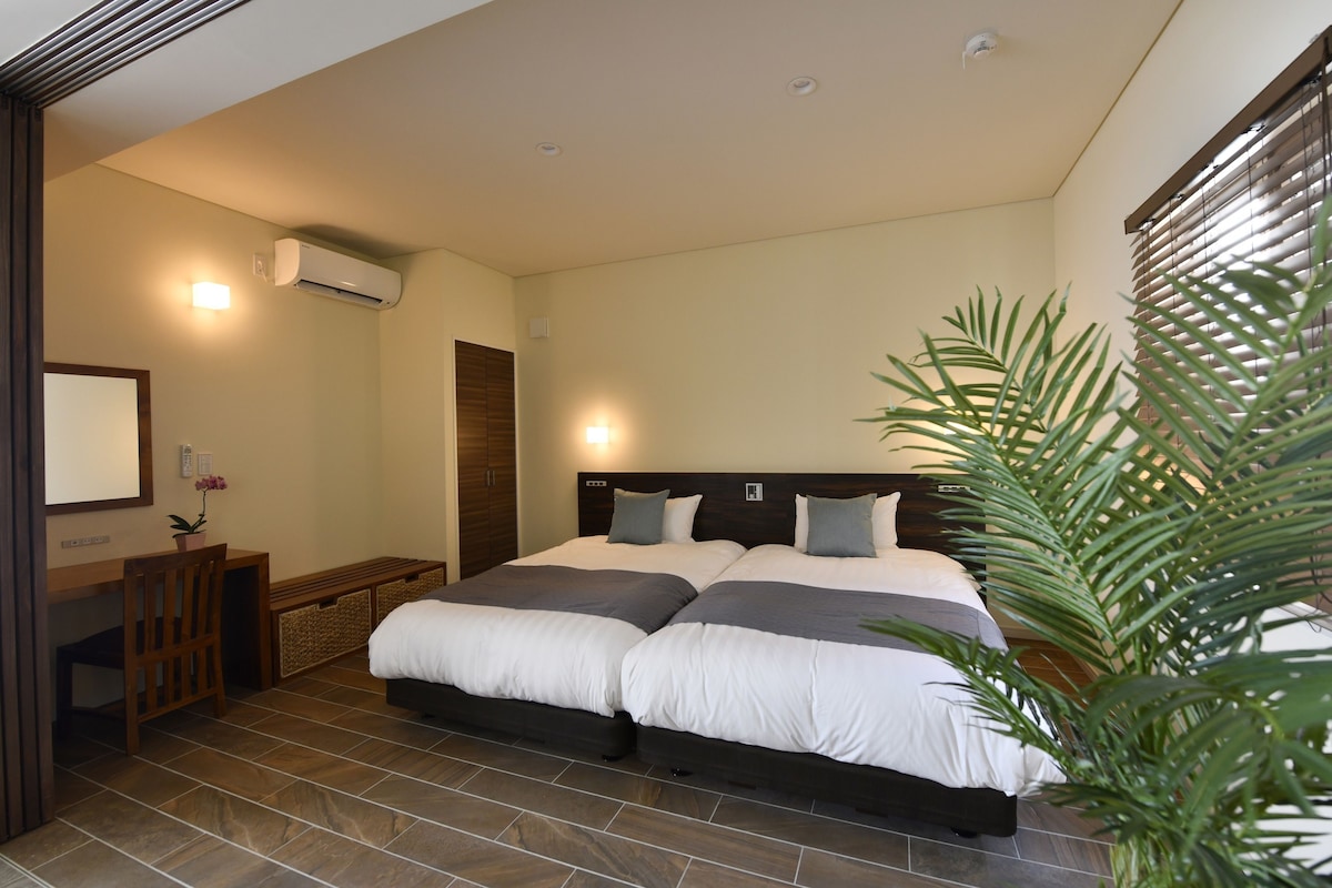Un dormitorio con una cama grande, un escritorio y una silla. Hay una planta en la esquina y un aire acondicionado en la pared.