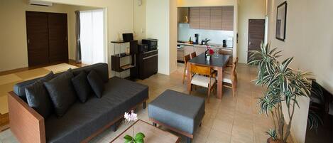 Una sala de estar moderna con sofá, otomanas y una mesa de comedor con sillas. Se aprecia una cocina al fondo con armarios y electrodomésticos.