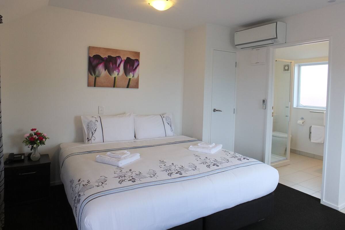 A hotel room with a bed, a painting of flowers, a nightstand, a vase with flowers, and a bathroom visible through an open door.