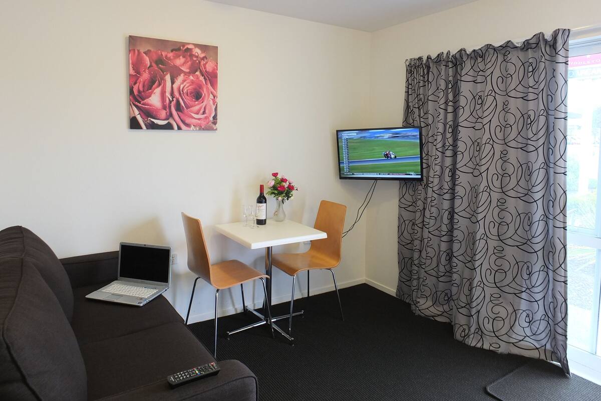 A hotel room with a sofa, a small table with a laptop, a TV, and a window with curtains.