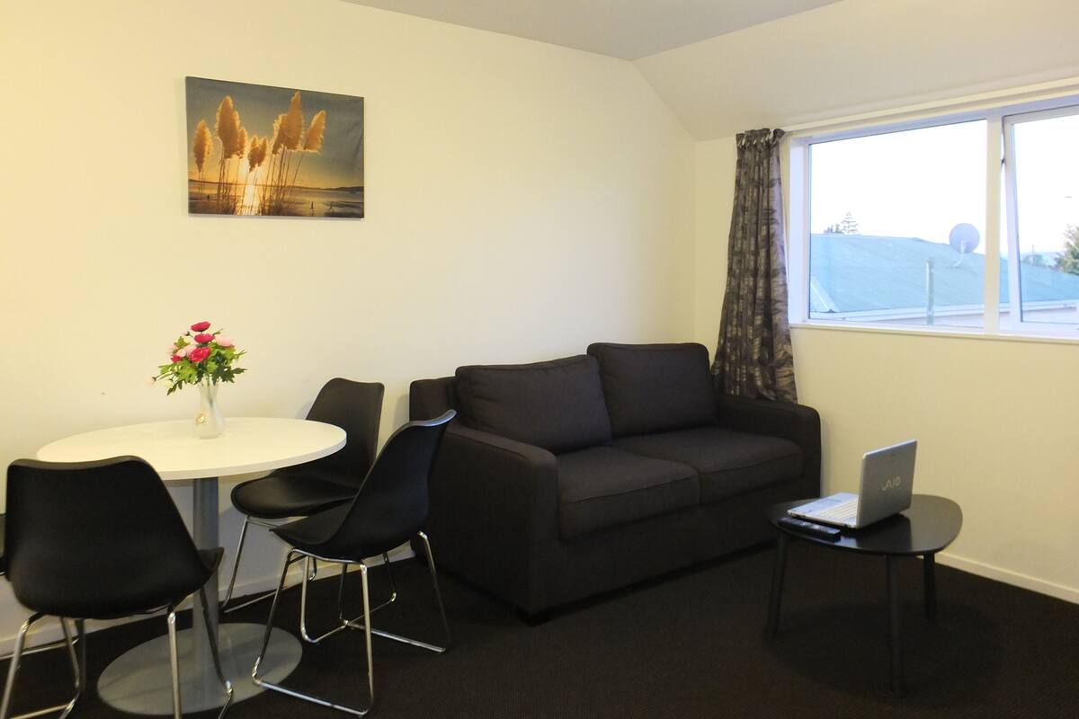 A modern living room with a black sofa, a round white table, two black chairs, a laptop on a small table, and a painting on the wall.
