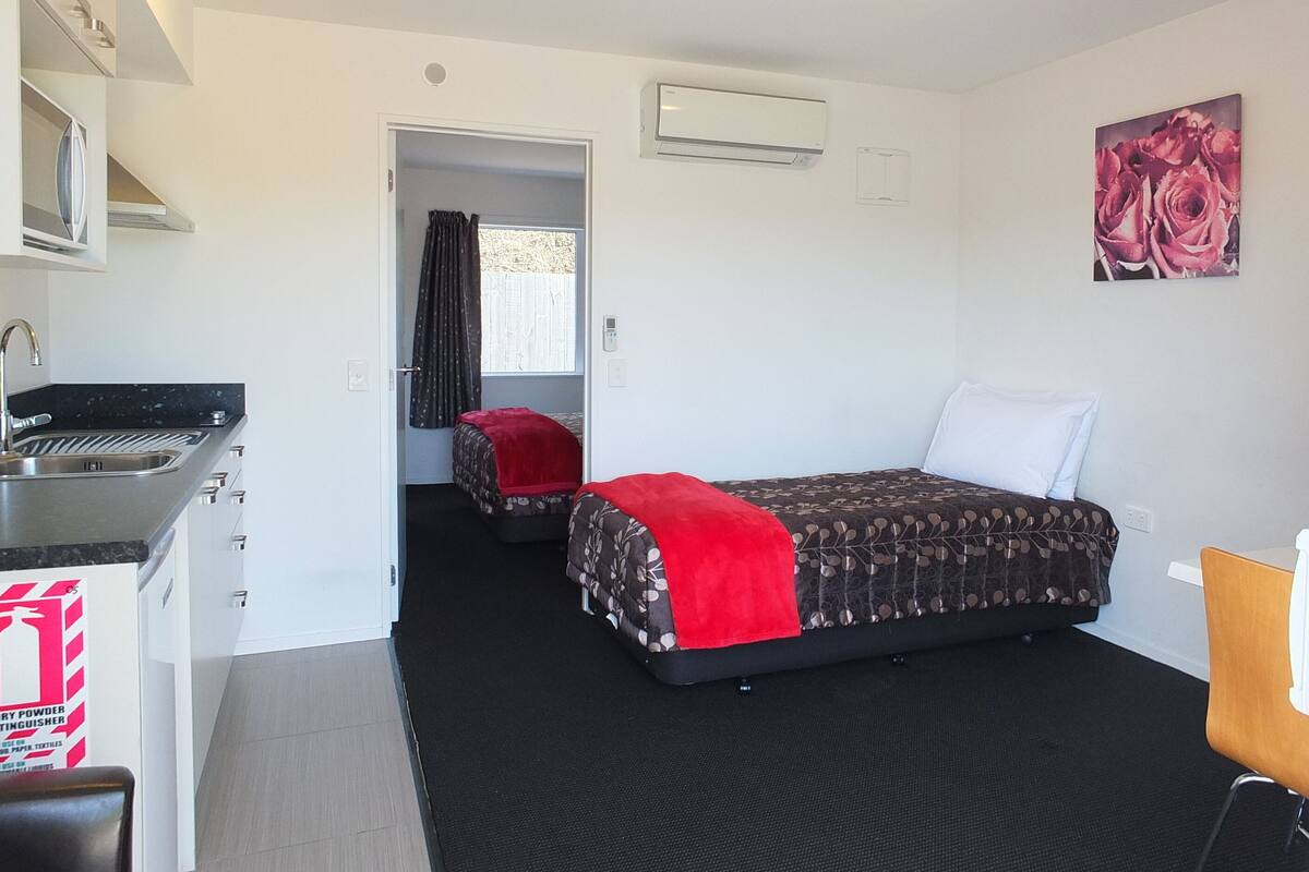 A compact hotel room with a kitchenette, a bed with a red blanket, a painting of roses, and a small table with a chair.