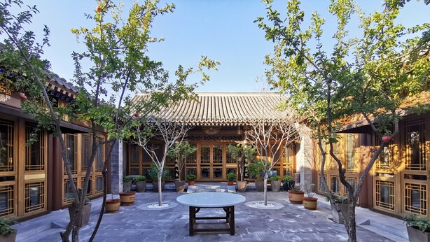 A courtyard with a central table, surrounded by potted plants and trees, with traditional wooden architecture in the background. - Cours Et Pavillons (Beijing)