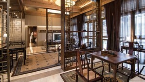 A spacious dining area with wooden tables and chairs, a decorative rug, and a view through a wooden partition to a kitchen area. - Cours Et Pavillons (Beijing)