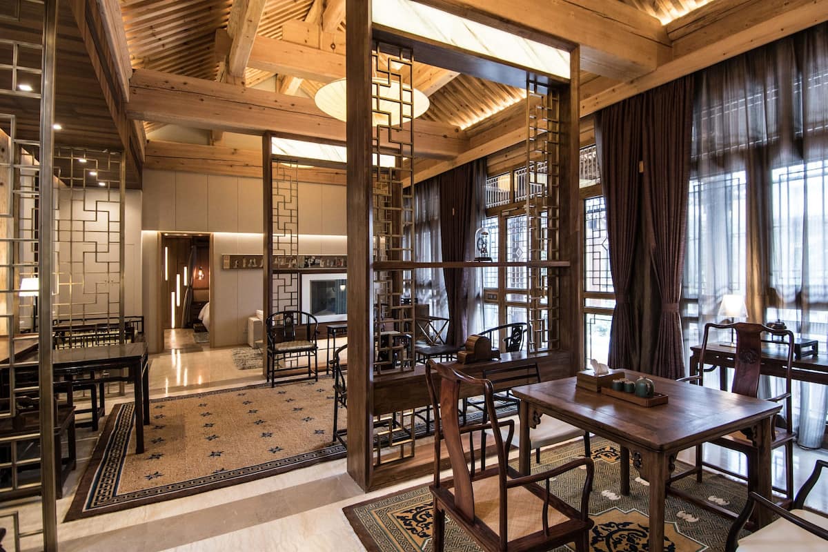 A spacious dining area with wooden tables and chairs, a decorative rug, and a view through a wooden partition to a kitchen area.