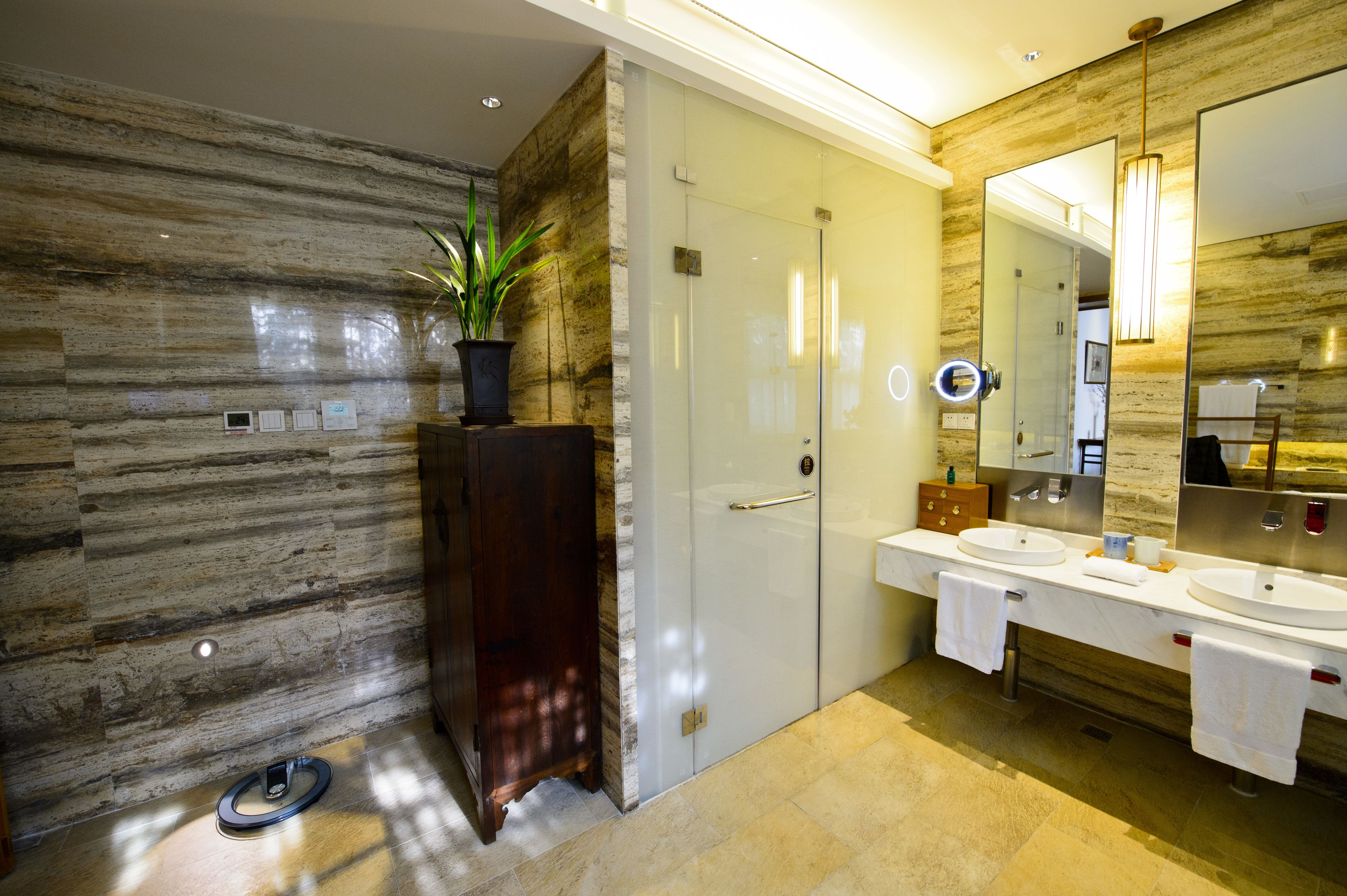 Une salle de bain moderne avec un mur en marbre, une plante en pot, un meuble en bois, une porte en verre, un grand miroir et un double vasque.