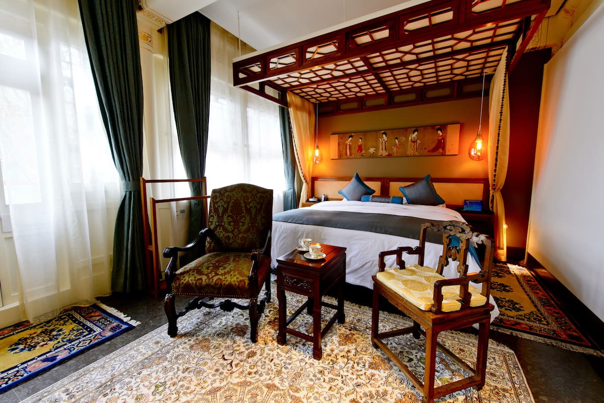 A hotel room with a bed, two chairs, a small table, and a decorative ceiling.