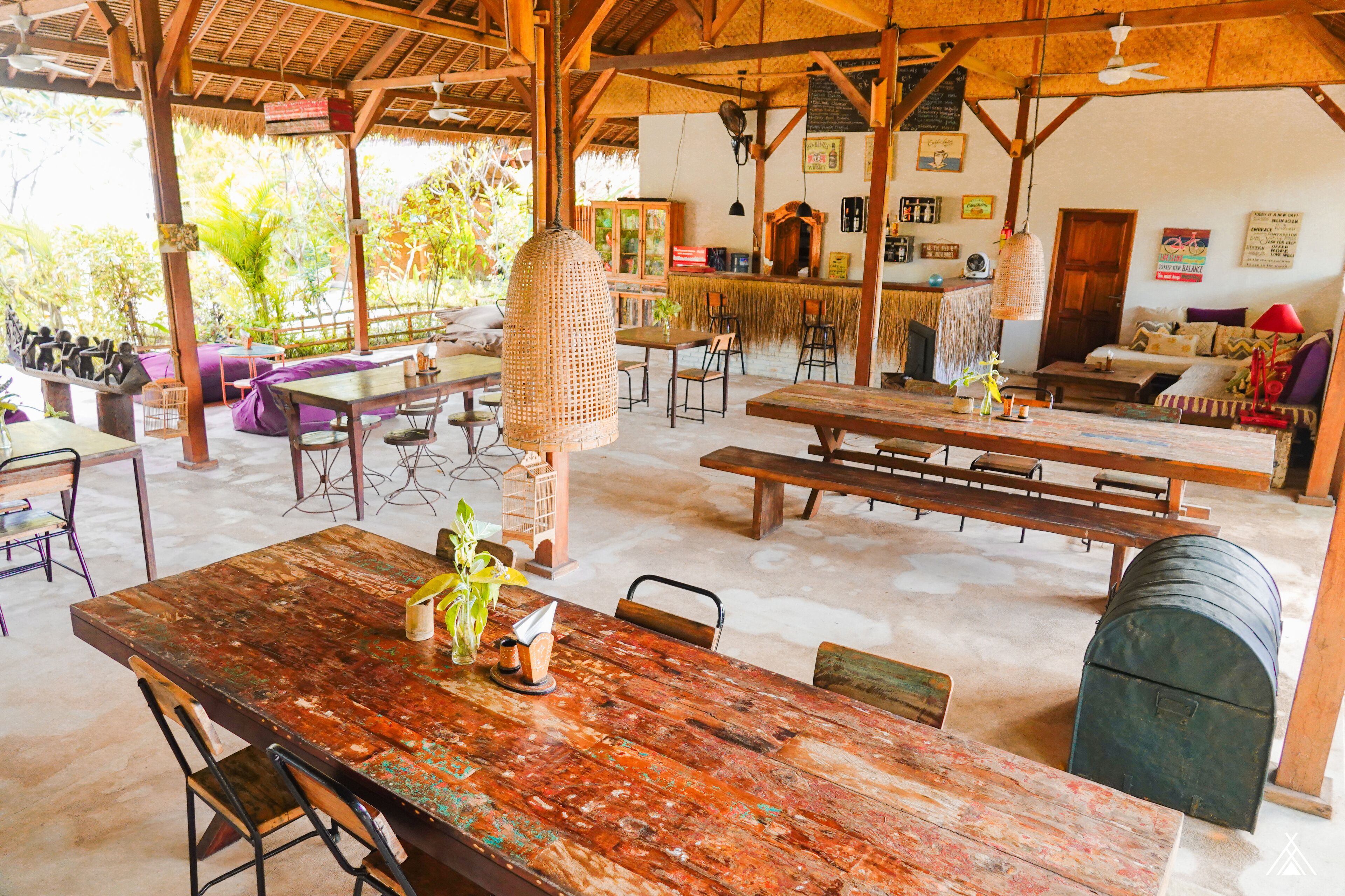 A rustic outdoor dining area with wooden tables and benches, surrounded by a thatched roof structure.