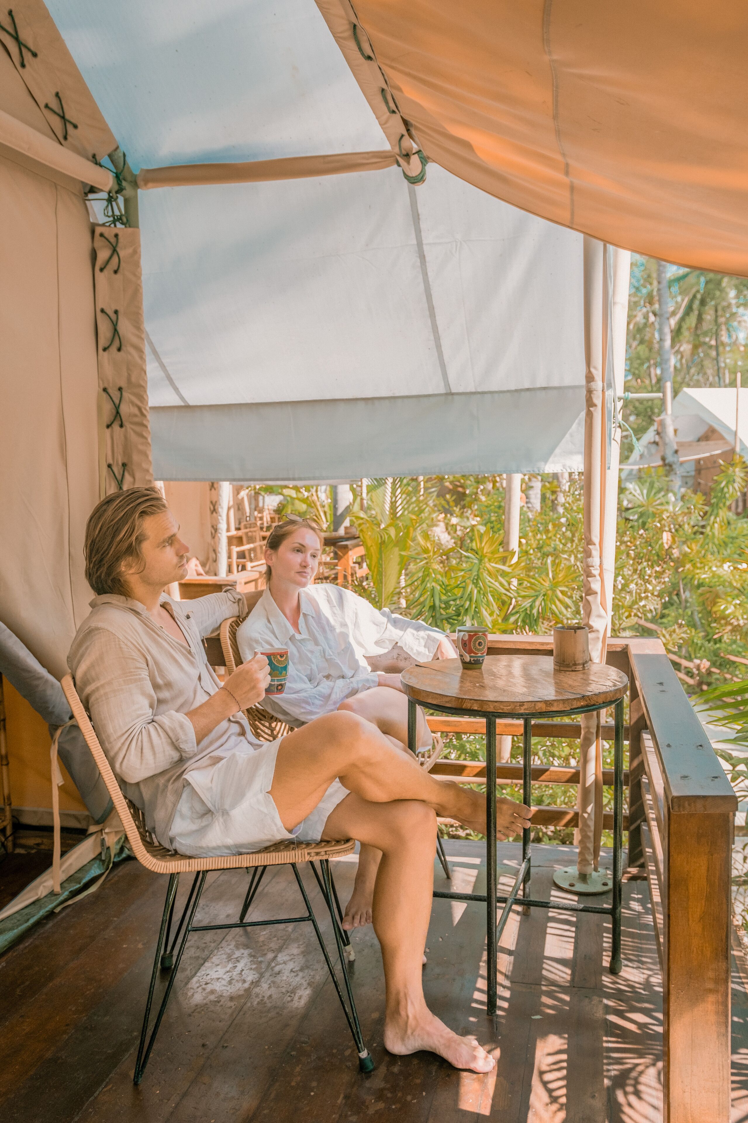A couple sitting on a wicker chair under a canopy, with a wooden table and a small potted plant between them.