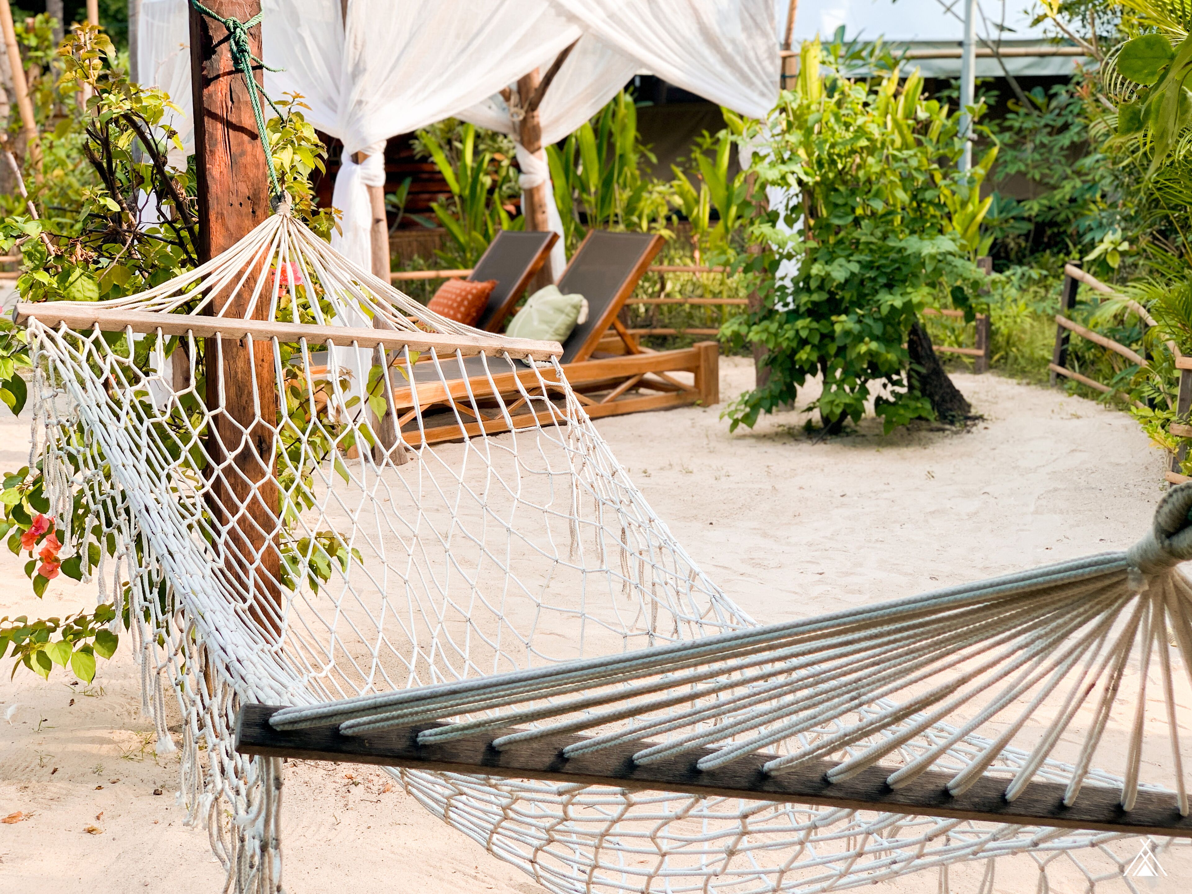 Un hamac et des chaises longues en bois, installés dans un jardin avec un sol sablonneux.