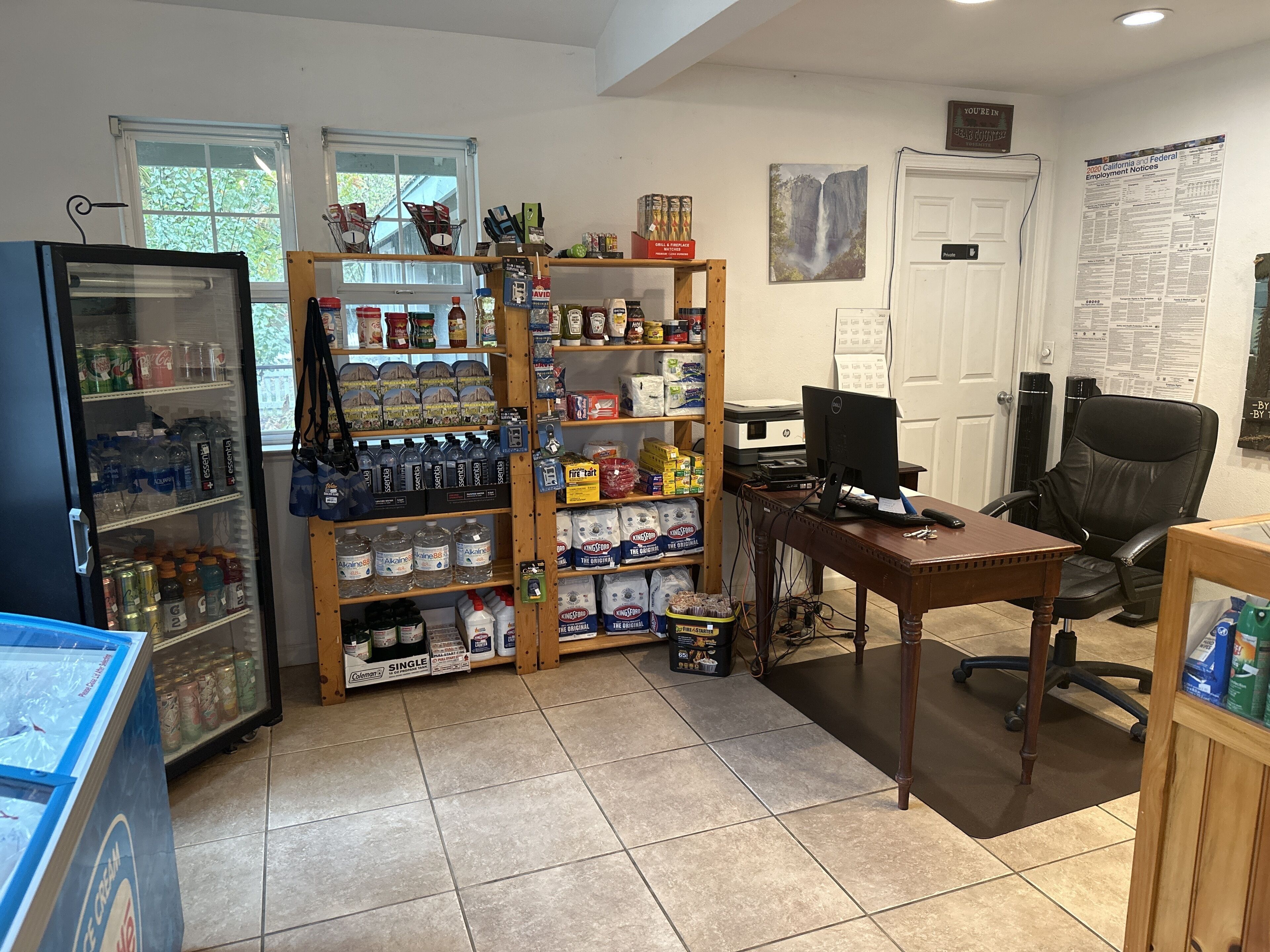 Una habitación con refrigerador lleno de bebidas, una estantería de madera con varios productos, un escritorio con computadora y una silla.