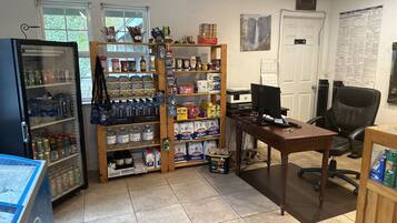 Una habitación con refrigerador lleno de bebidas, una estantería de madera con varios productos, un escritorio con computadora y una silla.