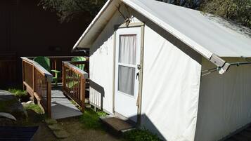 Une petite tente blanche, composée d’une seule pièce, avec une porte et une fenêtre, une terrasse en bois et une chaise verte.