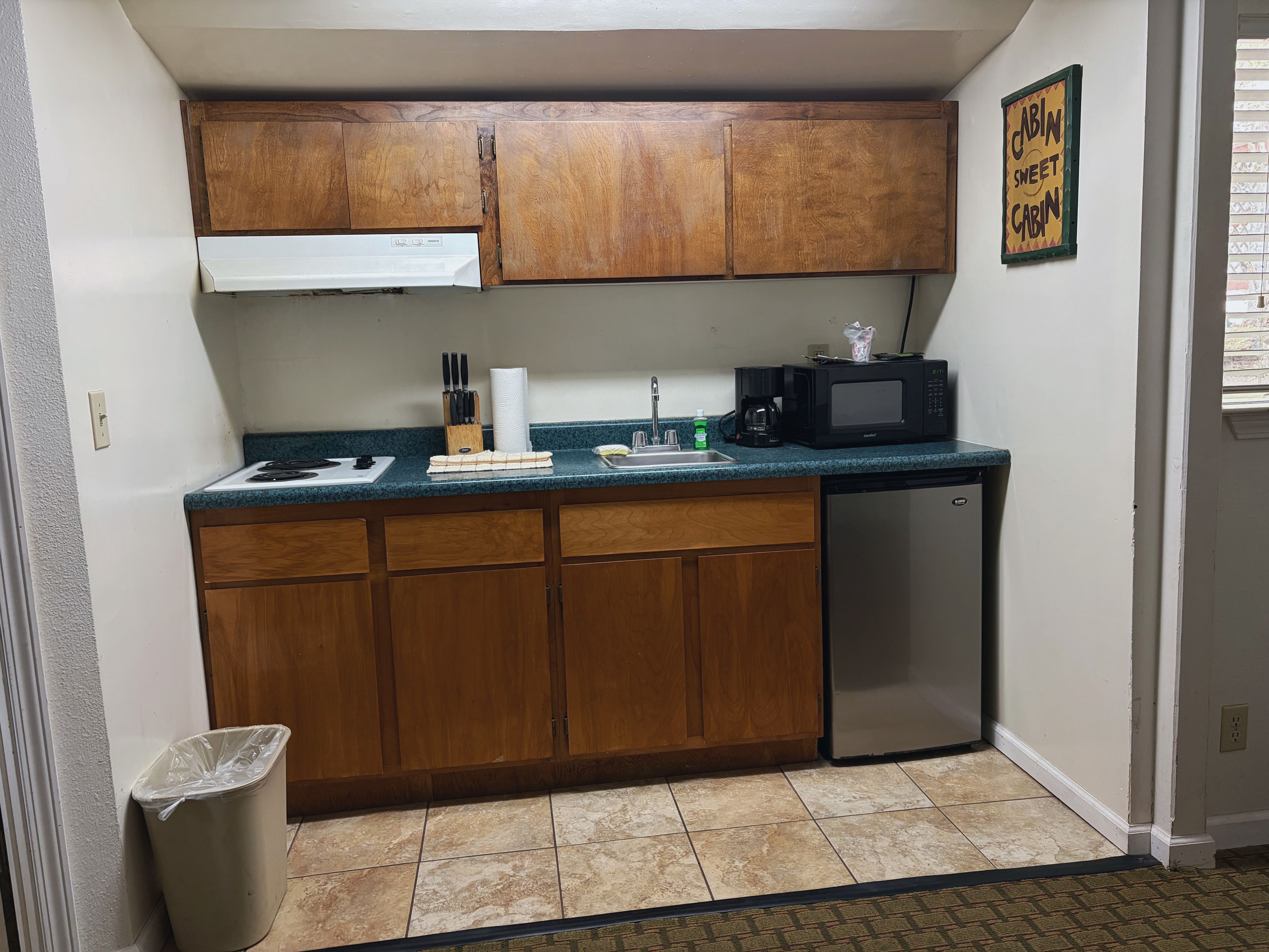 A compact kitchen with wooden cabinets, a microwave, a sink, and a trash bin.