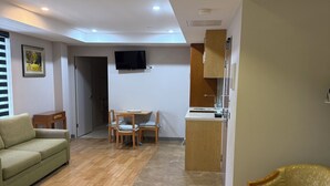 Una sala moderna con un sofá, una mesa de comedor con sillas, un televisor montado en la pared y una zona de cocina con armarios y un lavamanos.