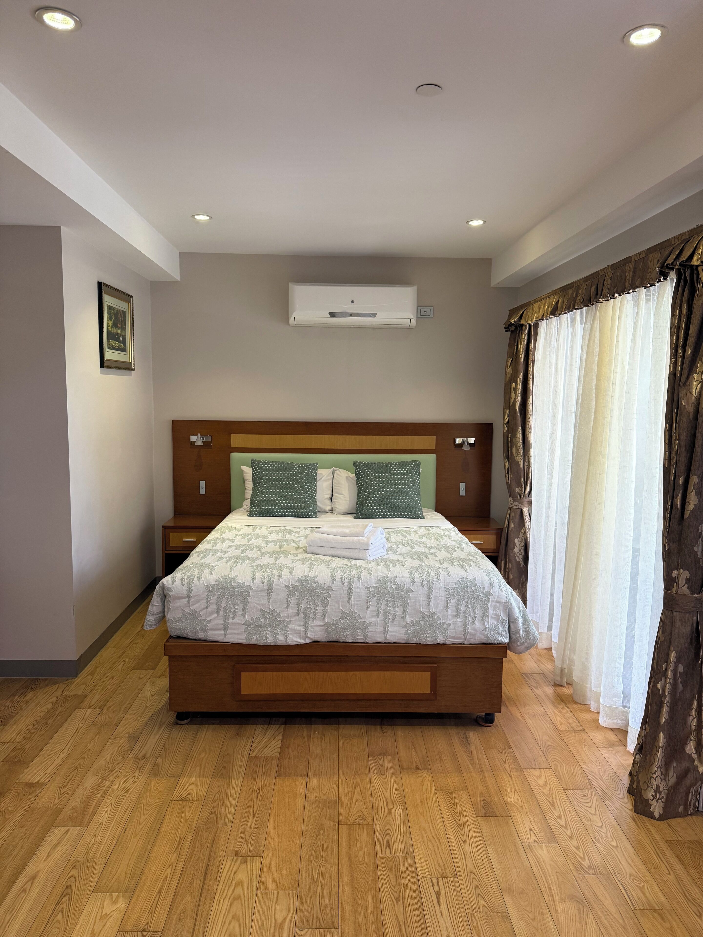 A bedroom with a wooden bed, a nightstand, a wall-mounted air conditioner, and a framed picture on the wall.