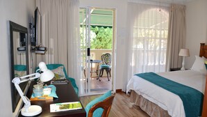 Habitación de hotel con cama, escritorio con lámpara, silla, televisor y puerta corrediza de vidrio que da a un área exterior con mesa y sillas.