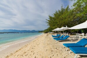 A beach with blue lounge chairs and white umbrellas, a sandy shore, and a mountain range in the background. - Danima Resort (Gili Trawangan)