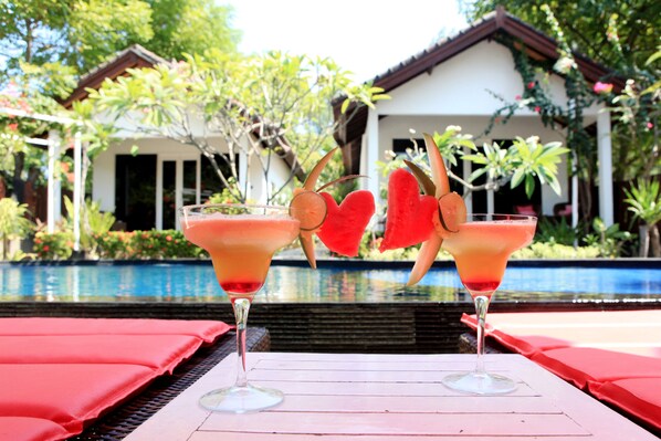 Two cocktails with heart-shaped garnishes on a table by a poolside, with a house and lush greenery in the background. - Danima Resort (Gili Trawangan)