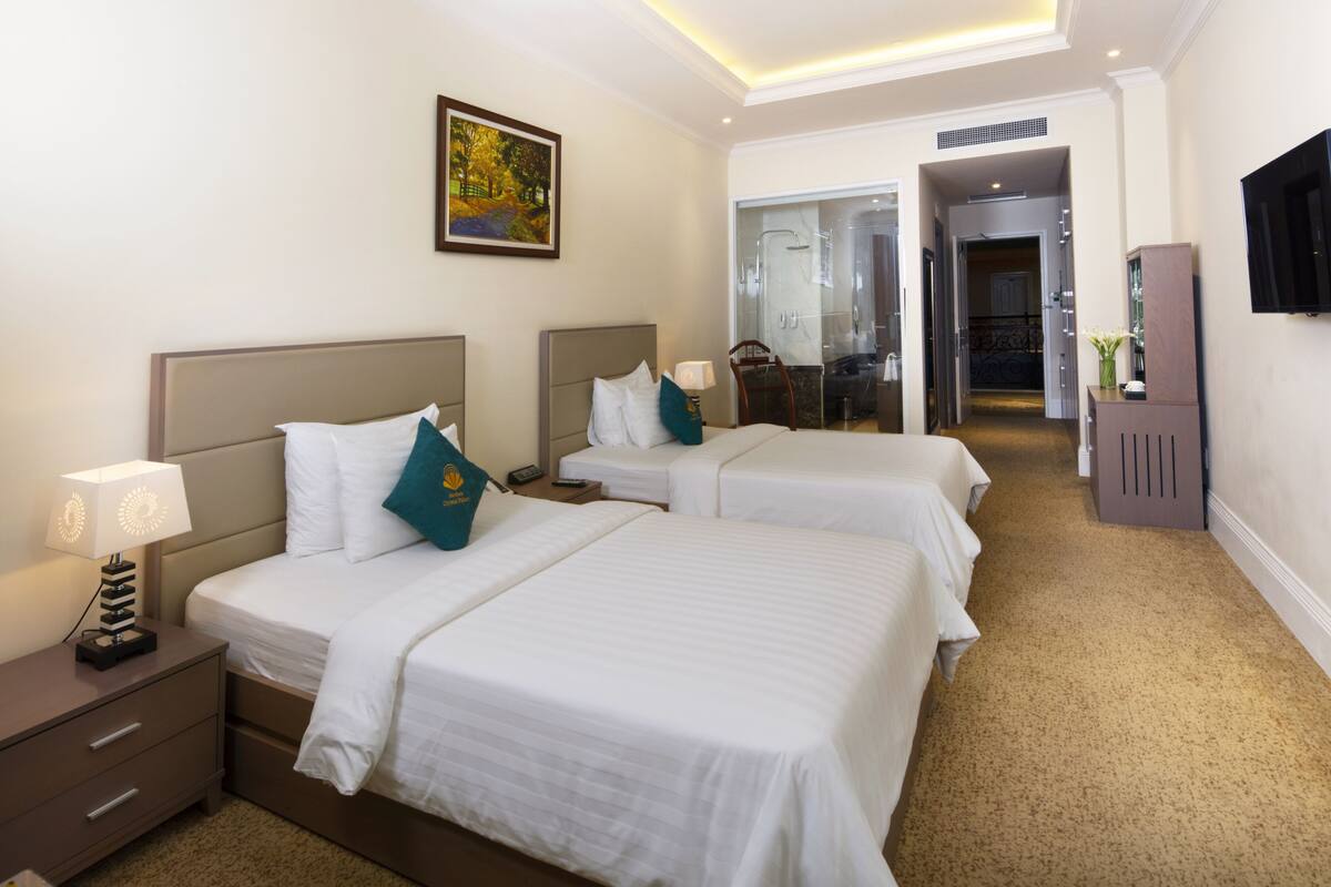 Habitación de hotel con dos camas, una mesita de noche, una lámpara, un cuadro, un televisor y un baño visible a través de una puerta abierta.