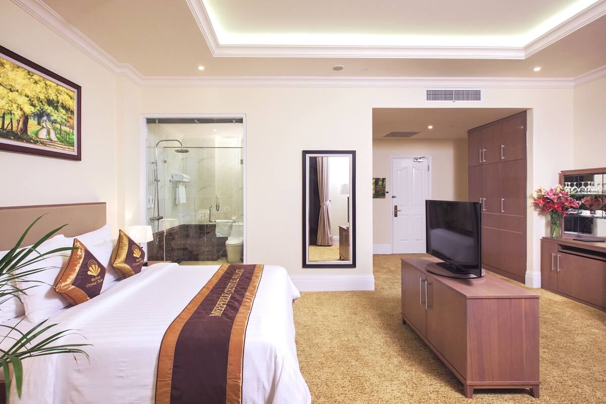 Habitación de hotel con una cama grande, televisor, armario y baño con ducha de vidrio, además de un cuadro en la pared.