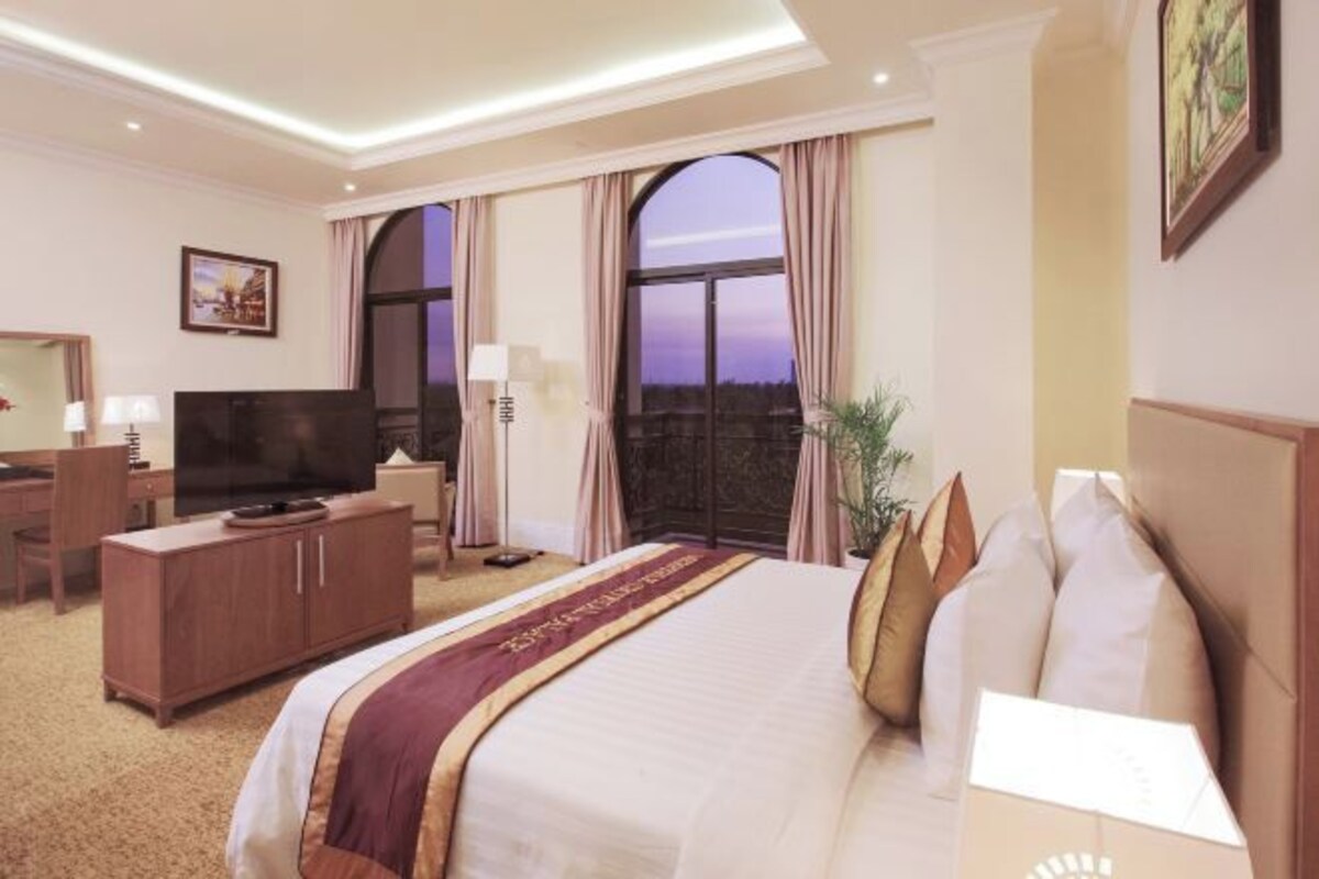 Quarto de hotel com uma cama grande, televisão, escrivaninha e varanda com cortinas.