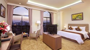 Ein Hotelzimmer mit einem großen Bett, einem Schreibtisch mit Computer, einem Sessel und einem Fernseher. Es gibt große Fenster mit Vorhängen, ein Bild an der Wand und Blick auf die Stadtlandschaft.