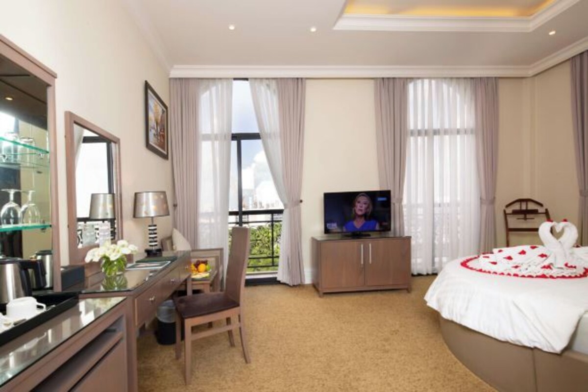 Ett hotellrum med en säng, en tv, ett skrivbord med en stol, ett litet bord med en vas med blommor och en balkong med gardiner.