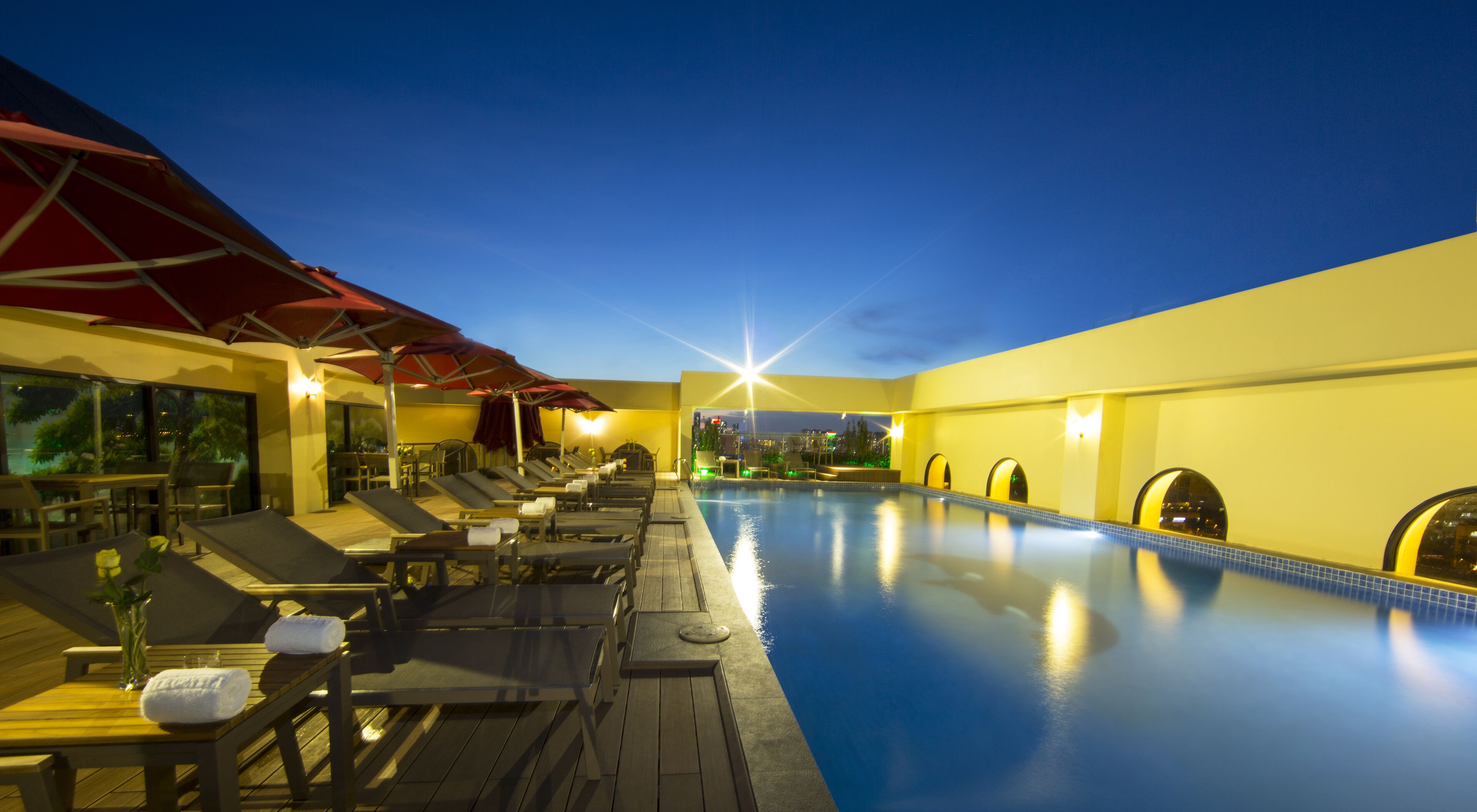 A poolside area with sun loungers and umbrellas, a clear sky, and a view of the city skyline.