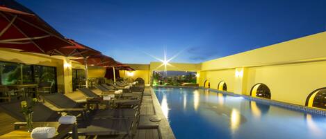 A poolside area with sun loungers and umbrellas, a clear sky, and a view of the city skyline.