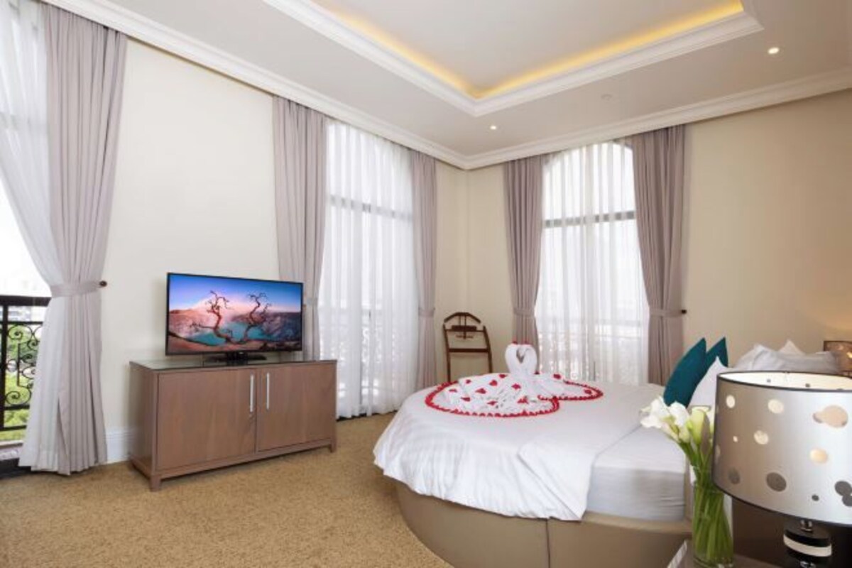 Quarto de hotel com uma cama, uma televisão sobre um móvel de madeira e um vaso com flores.