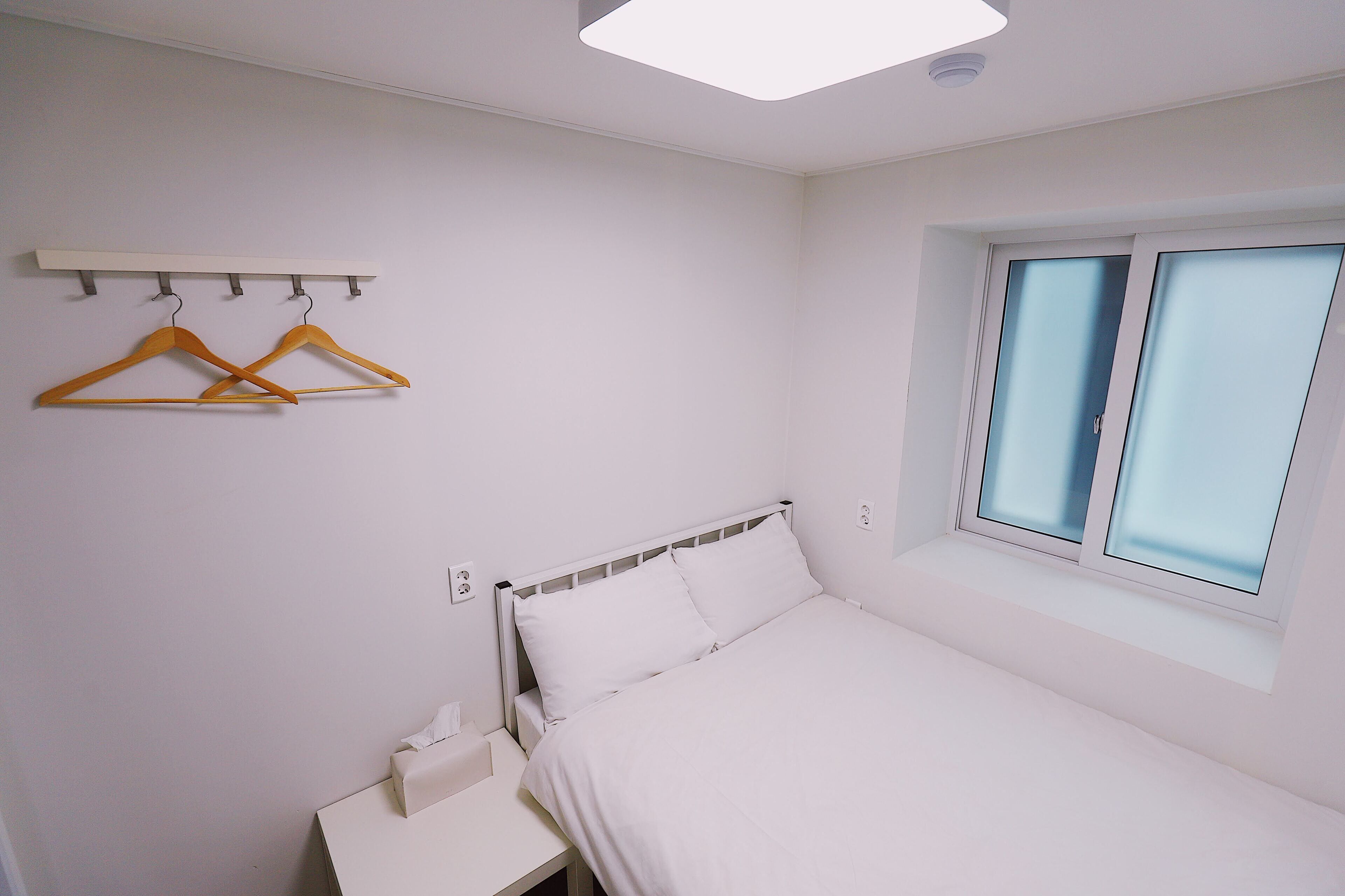A bedroom with a bed, a bedside table, a window, and a wall-mounted shelf with hangers.