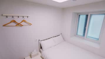 A bedroom with a bed, a bedside table, a window, and a wall-mounted shelf with hangers.