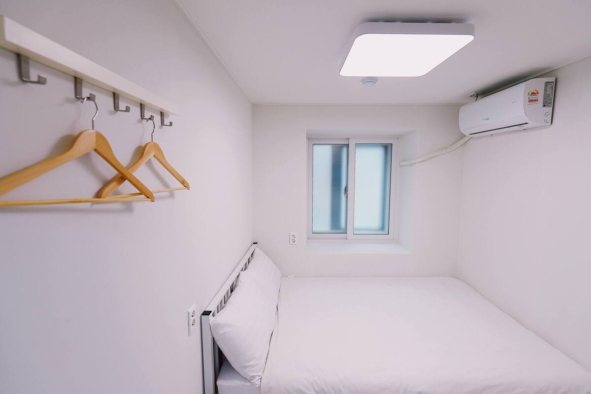 Una habitación pequeña y limpia con una cama, una ventana, aire acondicionado de pared y un perchero con perchas.