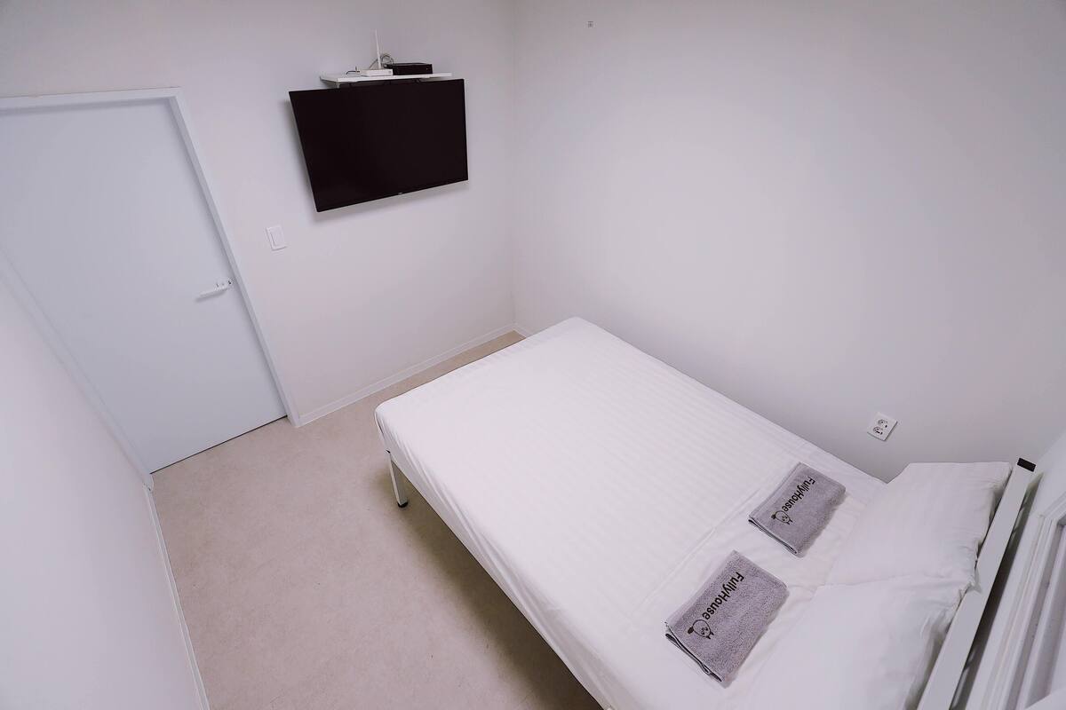 Una habitación pequeña y limpia con una cama individual, un televisor de pantalla plana en la pared y una puerta.