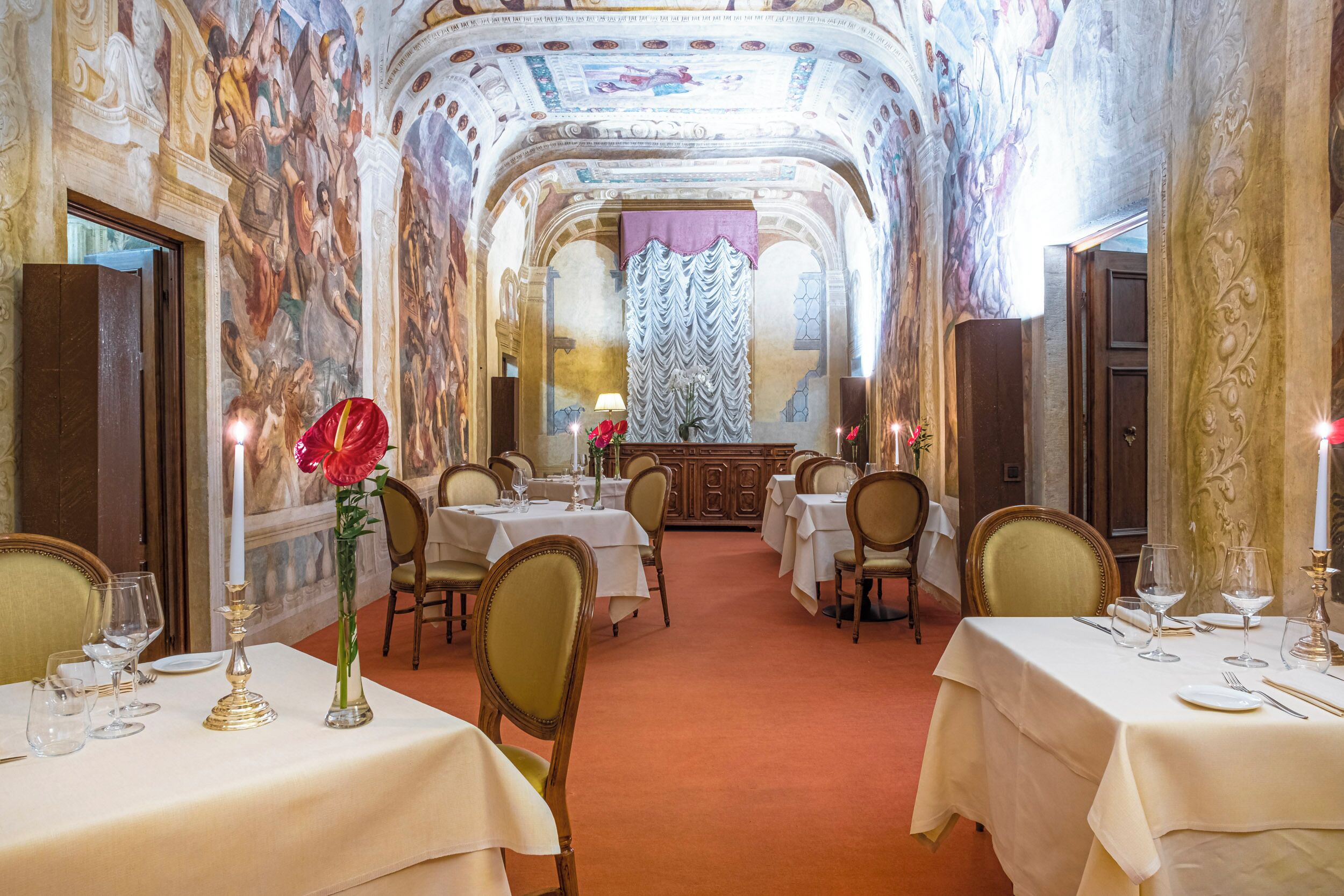 Un amplio comedor con frescos ornamentales en las paredes, varias mesas dispuestas para comer y una araña.