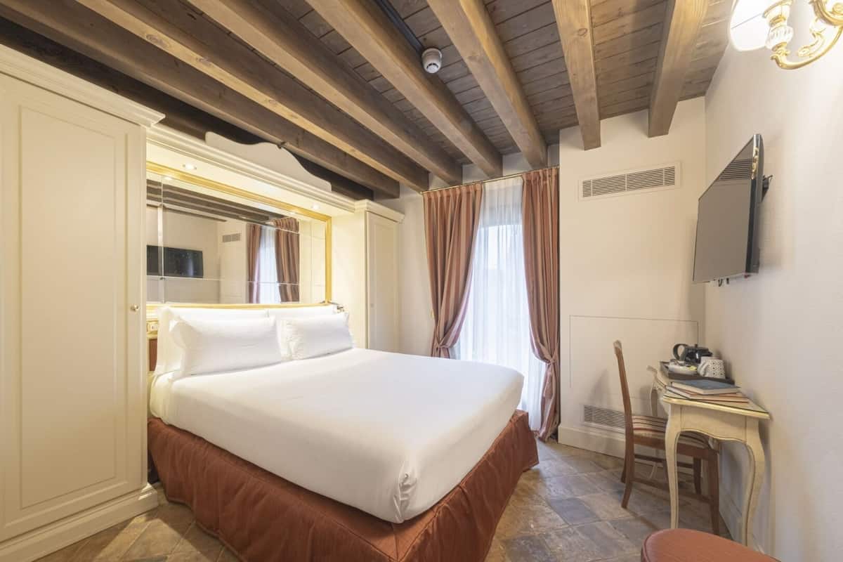 A hotel room with a large bed, a desk with a chair, a mirror, and a television on the wall.