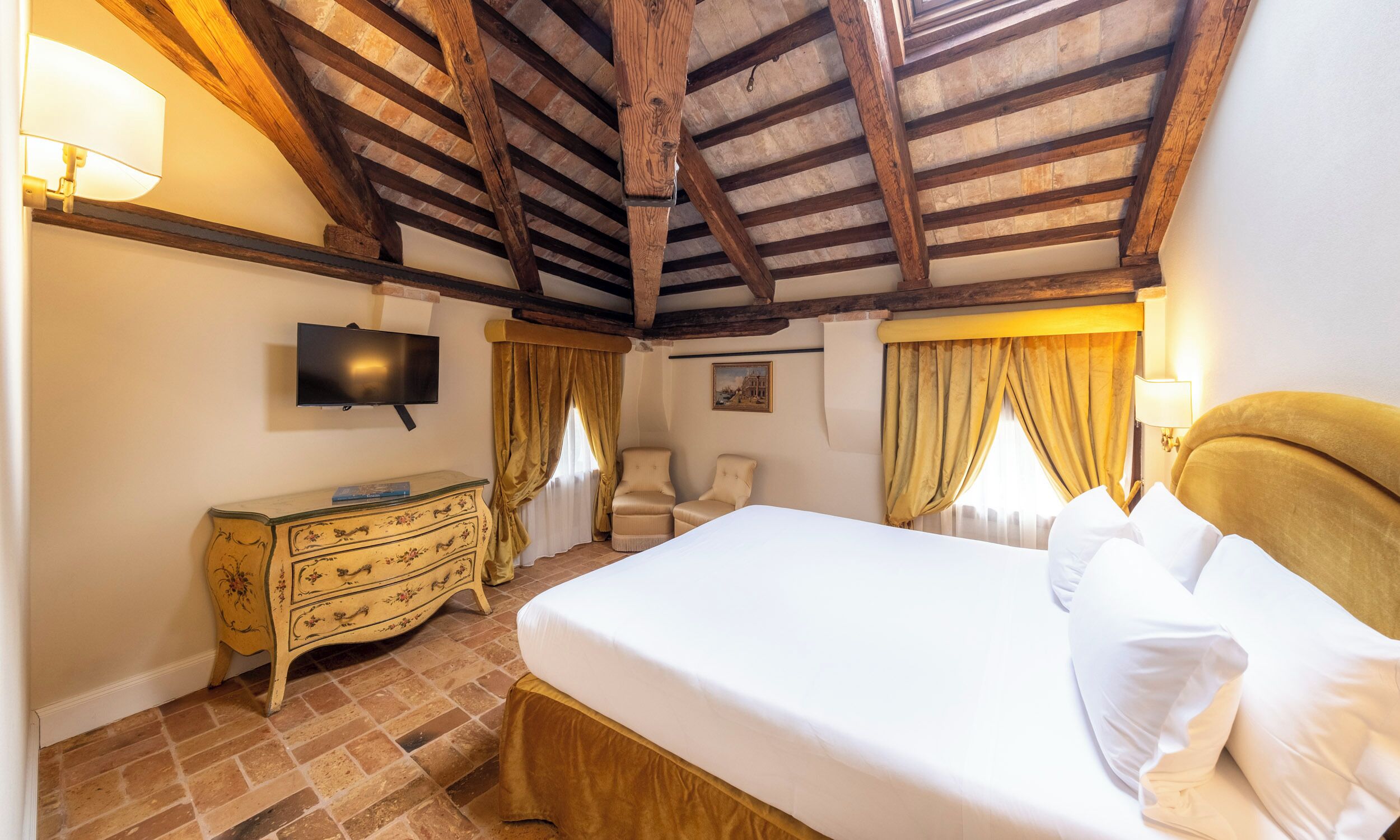 Ein Schlafzimmer mit Holzbalkendecke, einem Bett mit weißen Leinentüchern, einem an der Wand befestigten Fernseher, einem Schminktisch und zwei Stühlen.