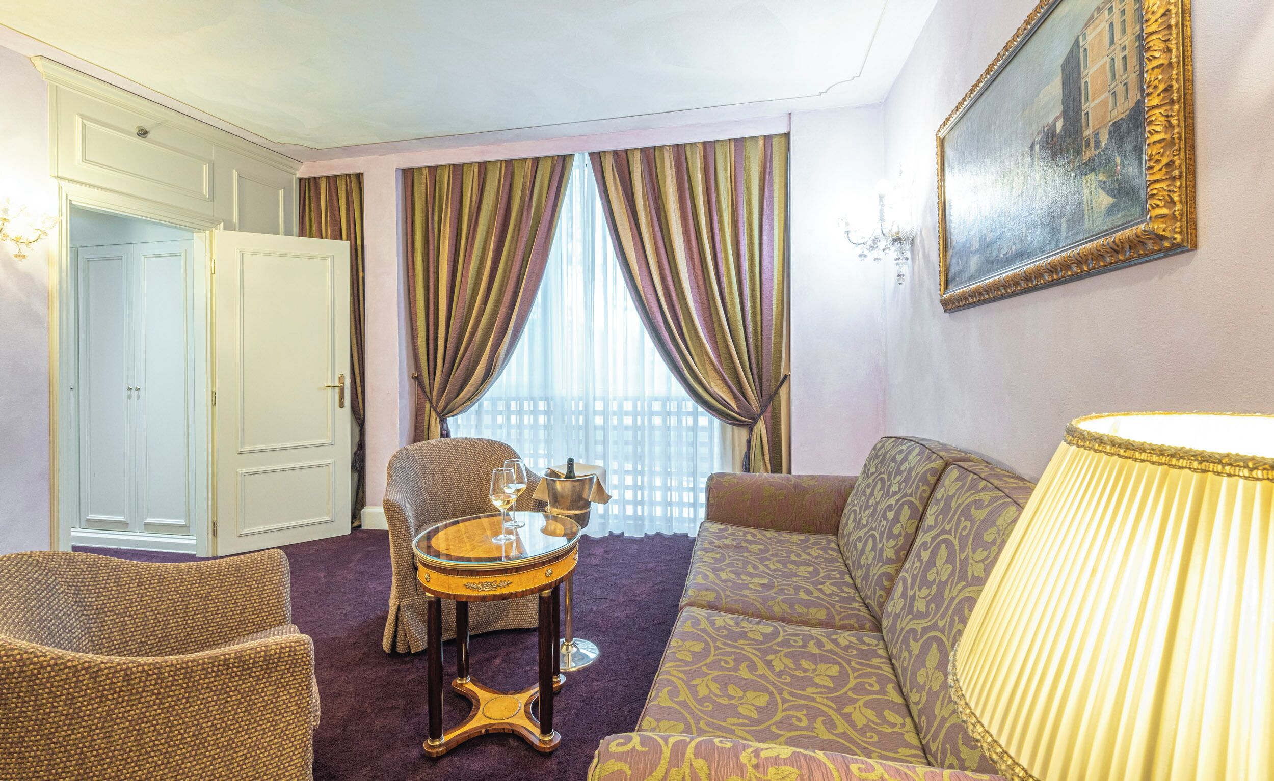 A hotel room with a sofa, armchair, a small round table with a lamp, and a window with curtains.