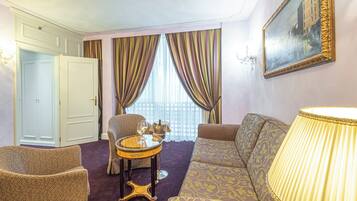 A hotel room with a sofa, armchair, a small round table with a lamp, and a window with curtains.