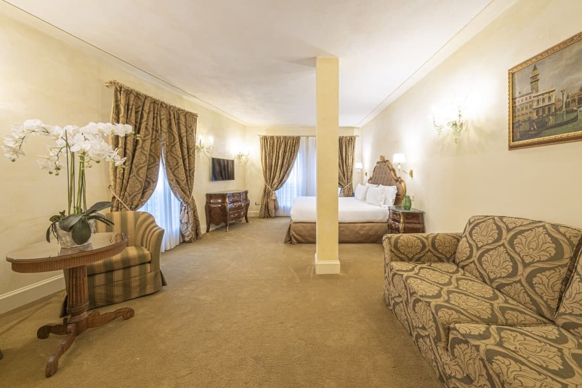 A hotel room with a large bed, a sofa, a small table with a vase of flowers, and a painting on the wall.